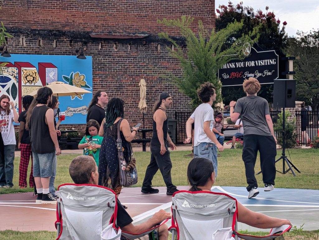 A group of people gather outdoors near mural and sign, with some sitting on lawn chairs and others standing or walking on the grass.