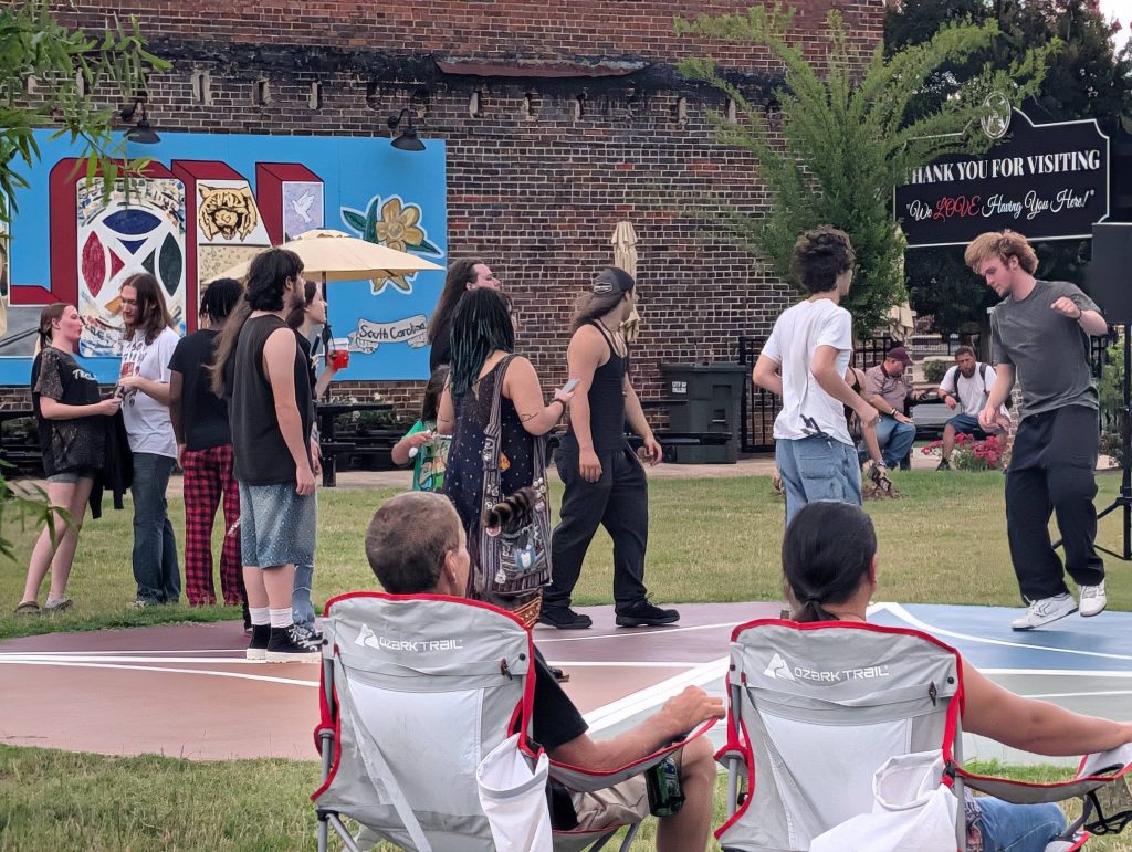 People standing and talking in a grassy area, some dancing, with two people sitting in chairs. A mural and a "Thank you for visiting" sign are visible.