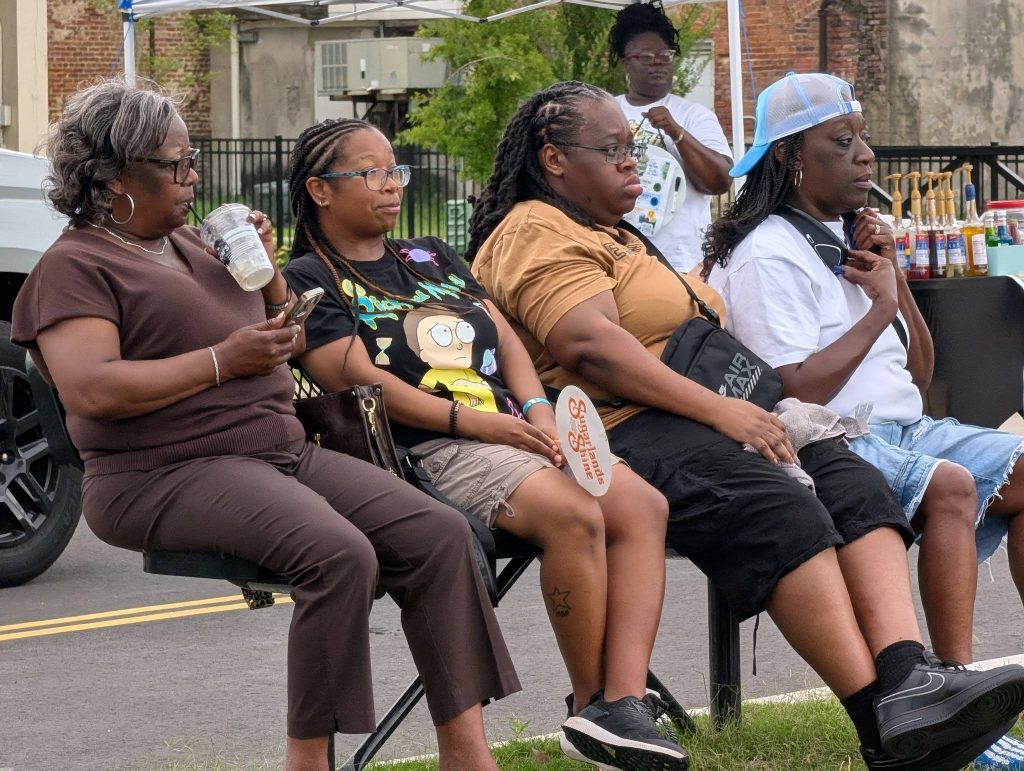 Four women sit in a row at an outdoor event, appearing relaxed. One drinks from a cup, others look ahead, with a vendor booth in the background.