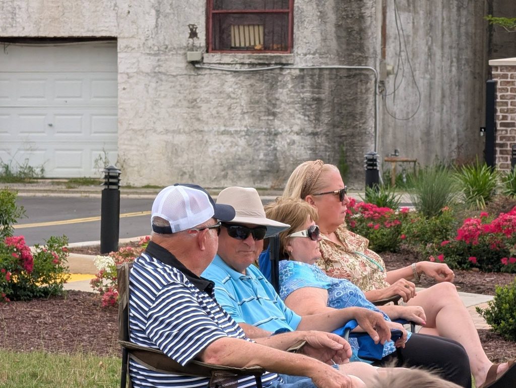 Four adults sit outdoors in lawn chairs near blooming flowers, wearing sunglasses and casual clothing, with a concrete building in the background.