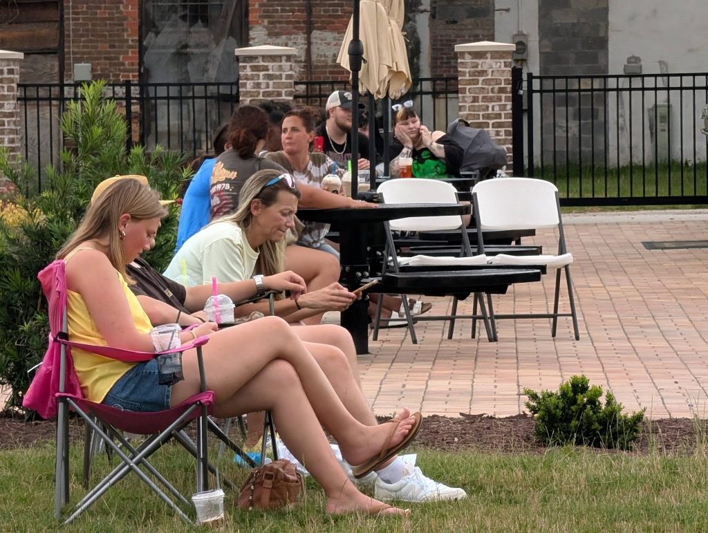 People sitting outdoors on lawn chairs and folding chairs, some drinking beverages, in a relaxed park area with plants and a brick walkway.