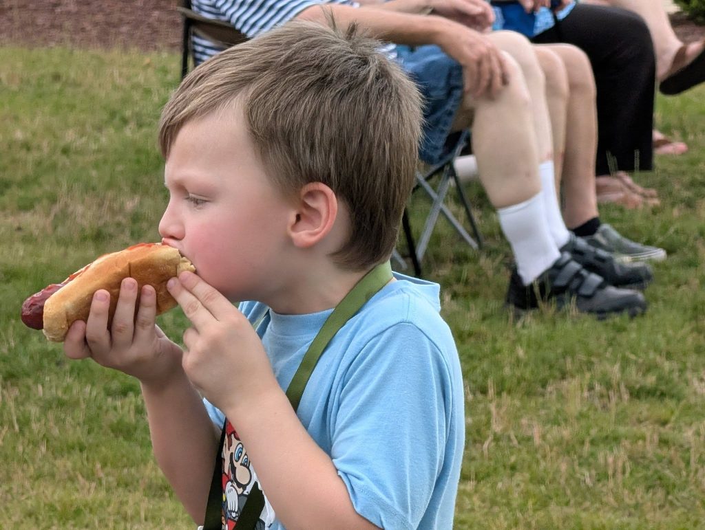 A young boy in a blue shirt eats a hot dog outdoors, with people sitting on chairs in the background.