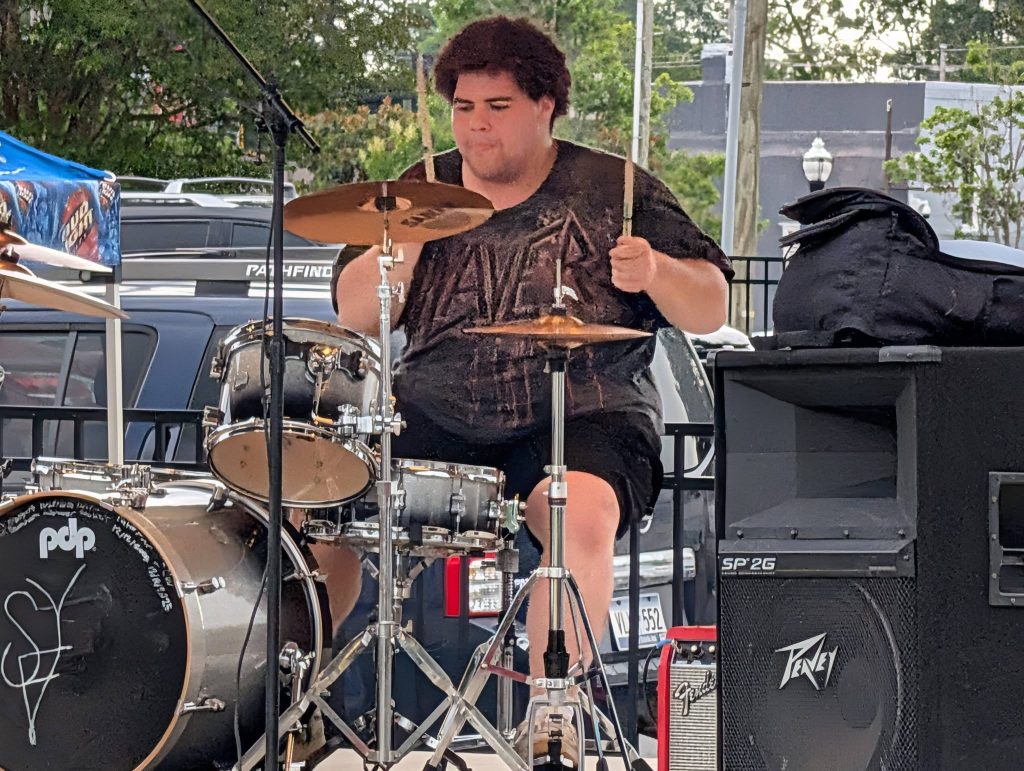A drummer wearing a black T-shirt performs on an outdoor stage surrounded by drum equipment, cars, and trees in the background.