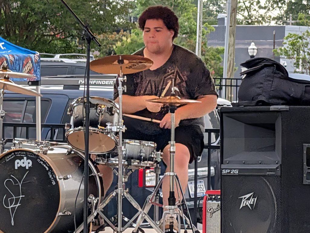 A person playing a drum set outdoors on a stage, with speakers, cars, and trees visible in the background.
