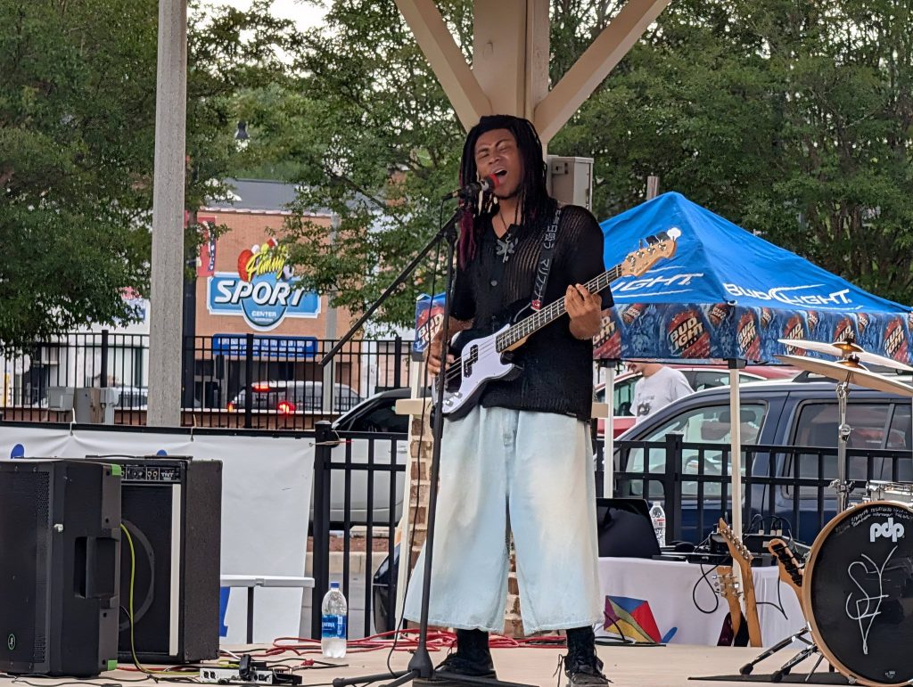 Musician with long hair playing electric bass guitar and singing on outdoor stage, with drum set and speakers, under a pavilion. Trees and a blue Bud Light tent in background.