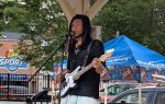 Musician performing outdoors under a pavilion, playing an electric bass guitar, singing into a microphone, with a blue Bud Light tent and trees in the background.