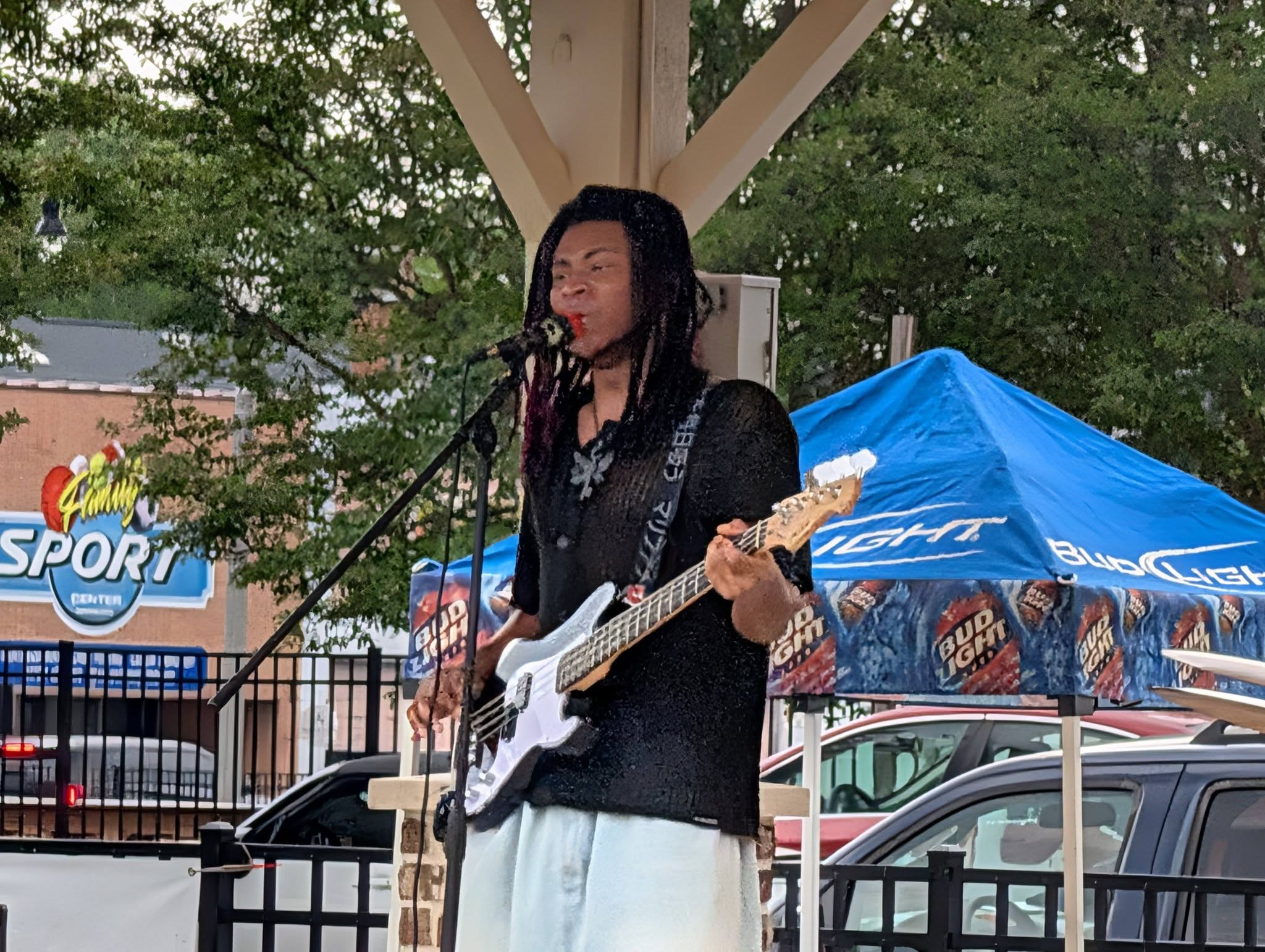 Musician performing outdoors under a pavilion, playing an electric bass guitar, singing into a microphone, with a blue Bud Light tent and trees in the background.