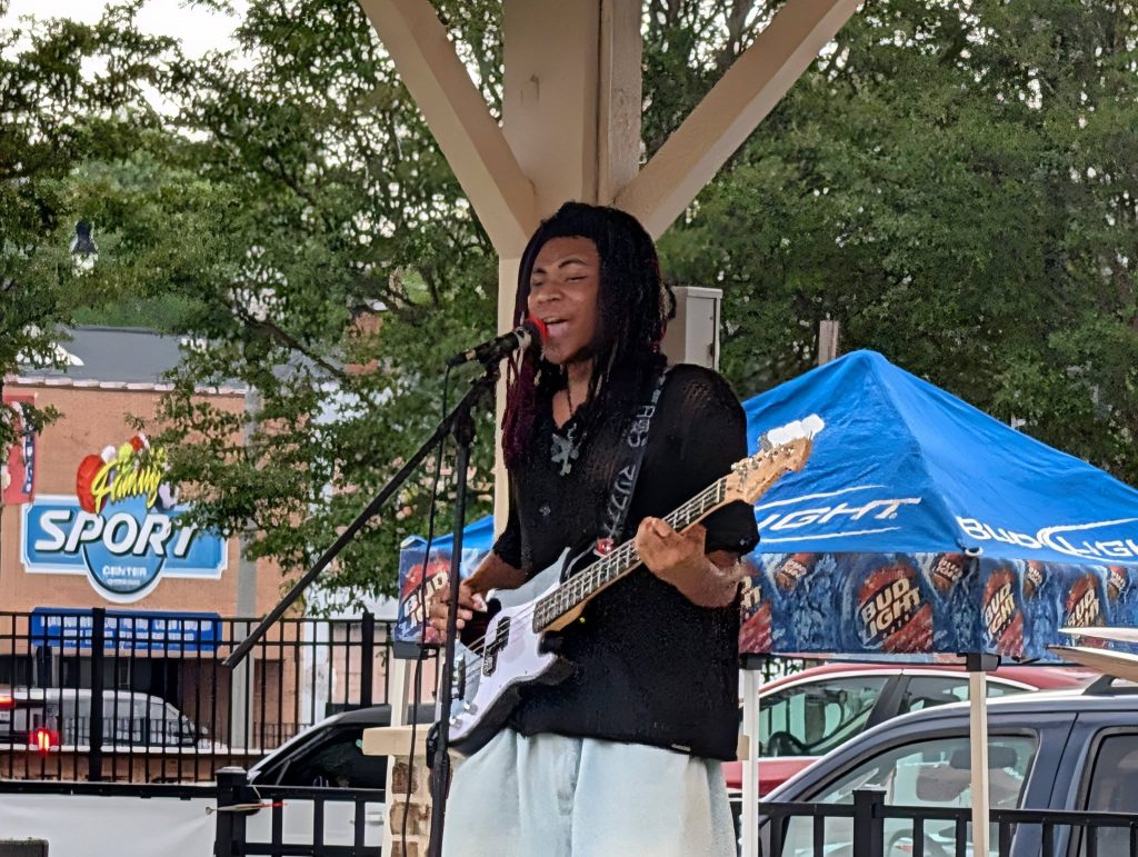 Musician performing outdoors on stage, playing bass guitar and singing into a microphone, with a blue Bud Light tent and cars in the background.