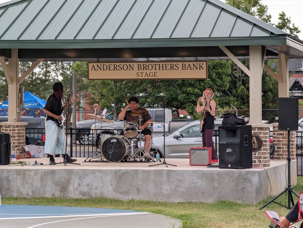 Three musicians performing on an outdoor stage under a pavilion with "Anderson Brothers Bank Stage" sign, featuring drums, guitars, and sound equipment.