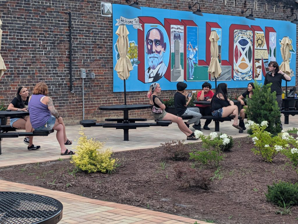 A group of people sit and talk at outdoor picnic tables near a mural that reads "DILLON" with images, against a brick wall backdrop.