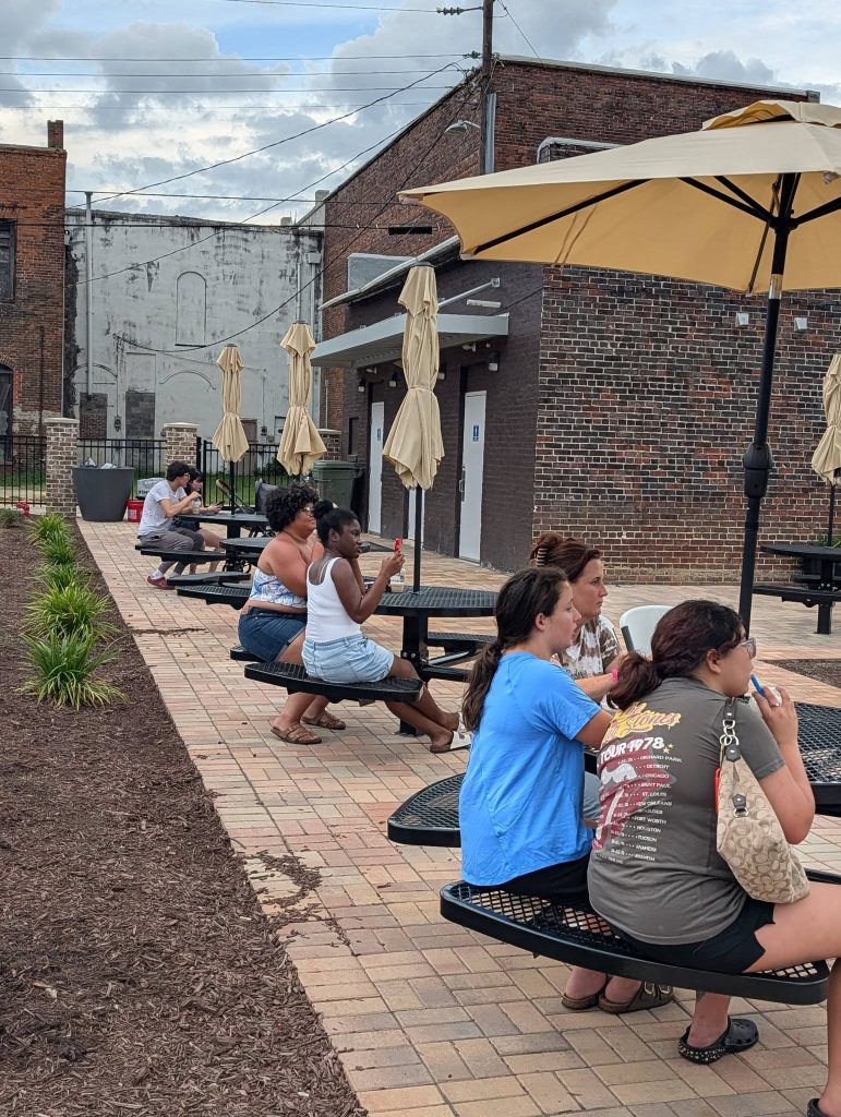People sitting at outdoor picnic tables, some eating or drinking, under large umbrellas near brick buildings on a cloudy day.