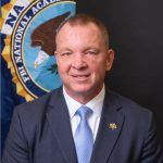 Man in a suit and tie smiling, seated in front of an FBI National Academy banner with an eagle emblem.