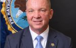 Man in a suit and tie smiling, seated in front of an FBI National Academy banner with an eagle emblem.