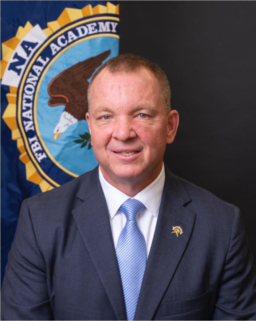 Man in a suit and tie smiling, seated in front of an FBI National Academy banner with an eagle emblem.