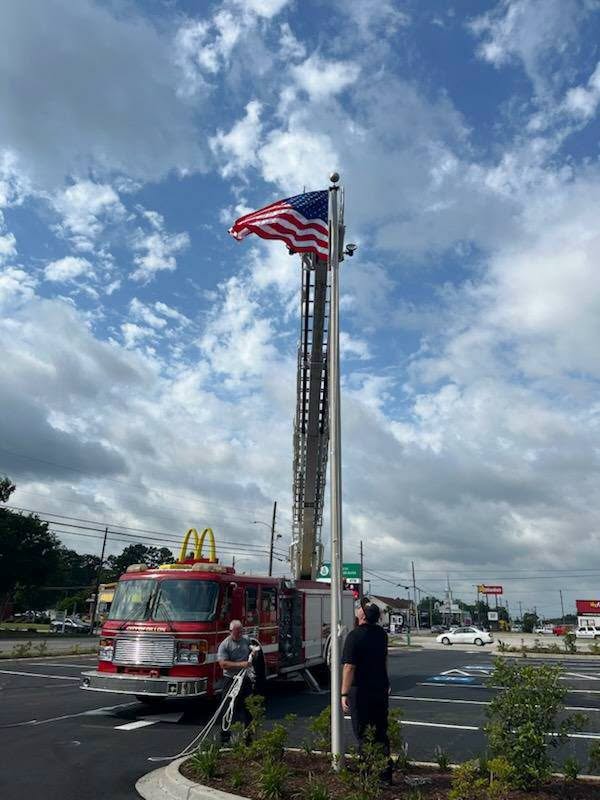 A fire truck raises an American flag on a pole in a parking lot, with people assisting and a McDonald's in the background under a partly cloudy sky.