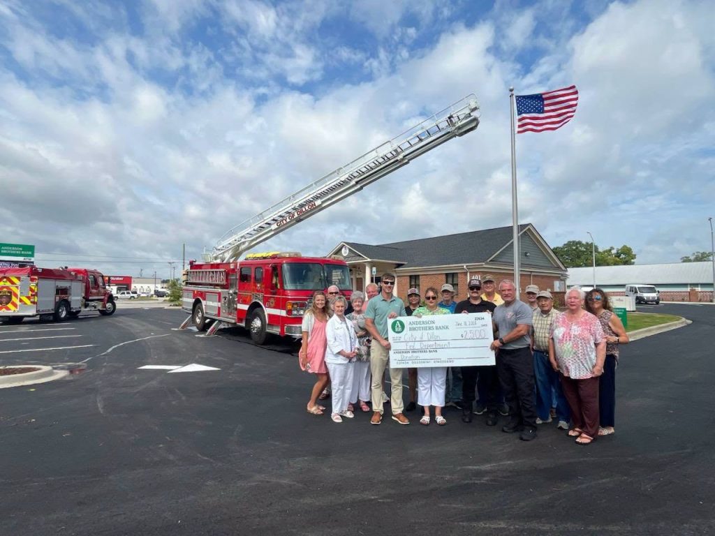A group of people holds a large check in front of fire trucks and an American flag outside a building on a sunny day.