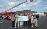 A group stands in front of a fire truck and American flag, holding a large check for $2,500 made out to the City of Dillon Fire Department.