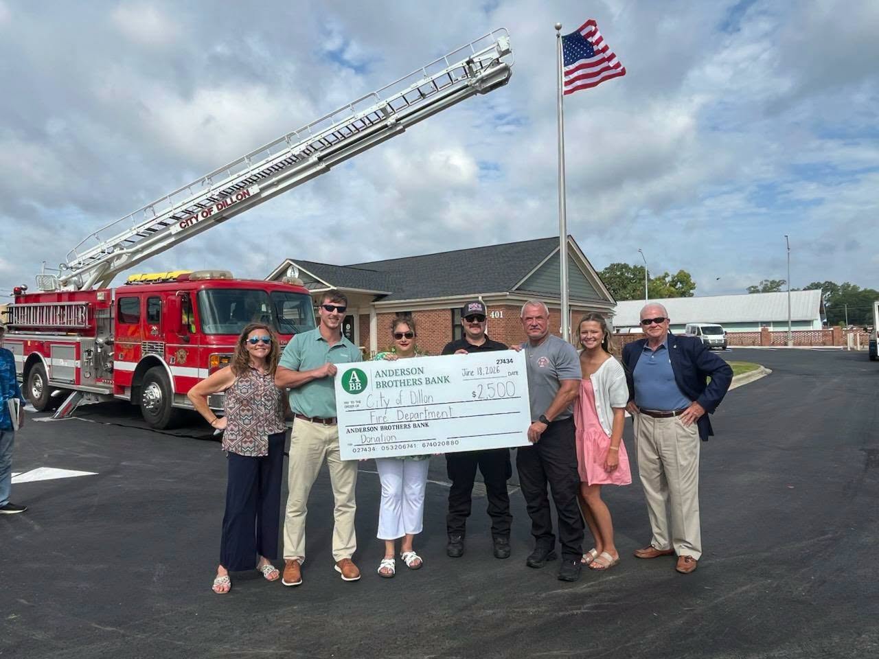 A group stands in front of a fire truck and American flag, holding a large check for $2,500 made out to the City of Dillon Fire Department.