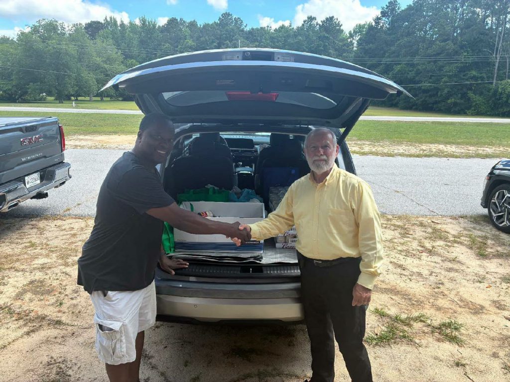 Two men shaking hands next to an open SUV trunk filled with supplies, in an outdoor parking area with trees and grass in the background.