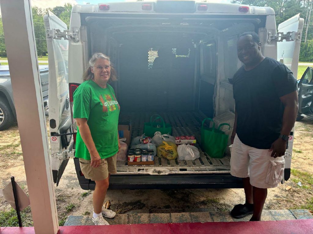 Two people stand by an open van filled with bags of groceries and canned goods, smiling outdoors on a sunny day.