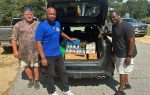 Three men stand next to an open car trunk filled with canned goods and food donations on a sunny day.