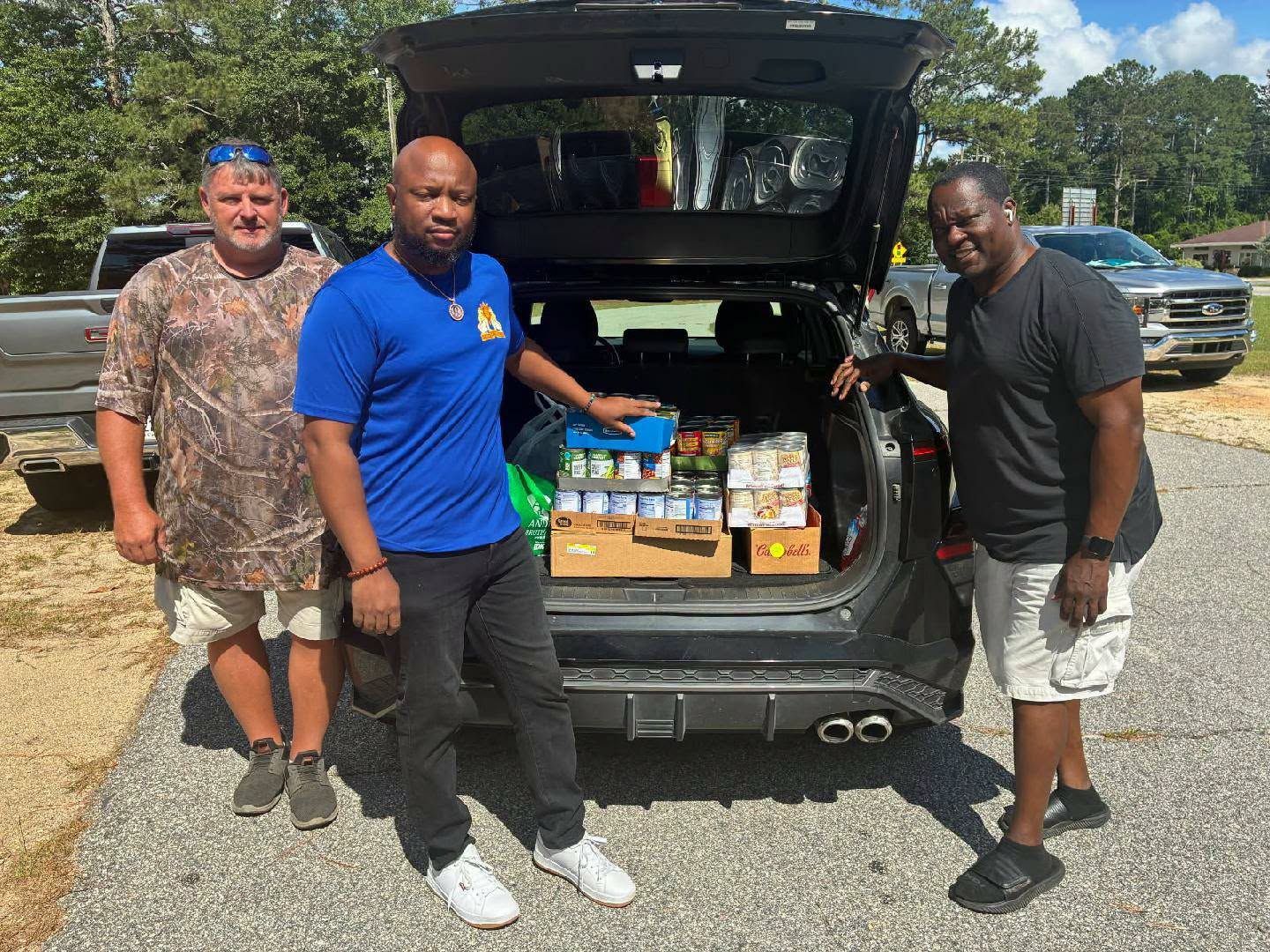Three men stand next to an open car trunk filled with canned goods and food donations on a sunny day.