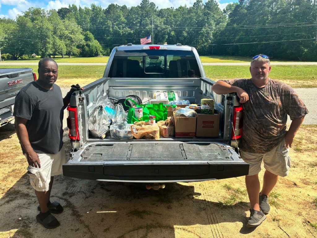 Two men stand by a pickup truck with its tailgate down, displaying bags and boxes of groceries and supplies in the truck bed. Trees and grass in the background.