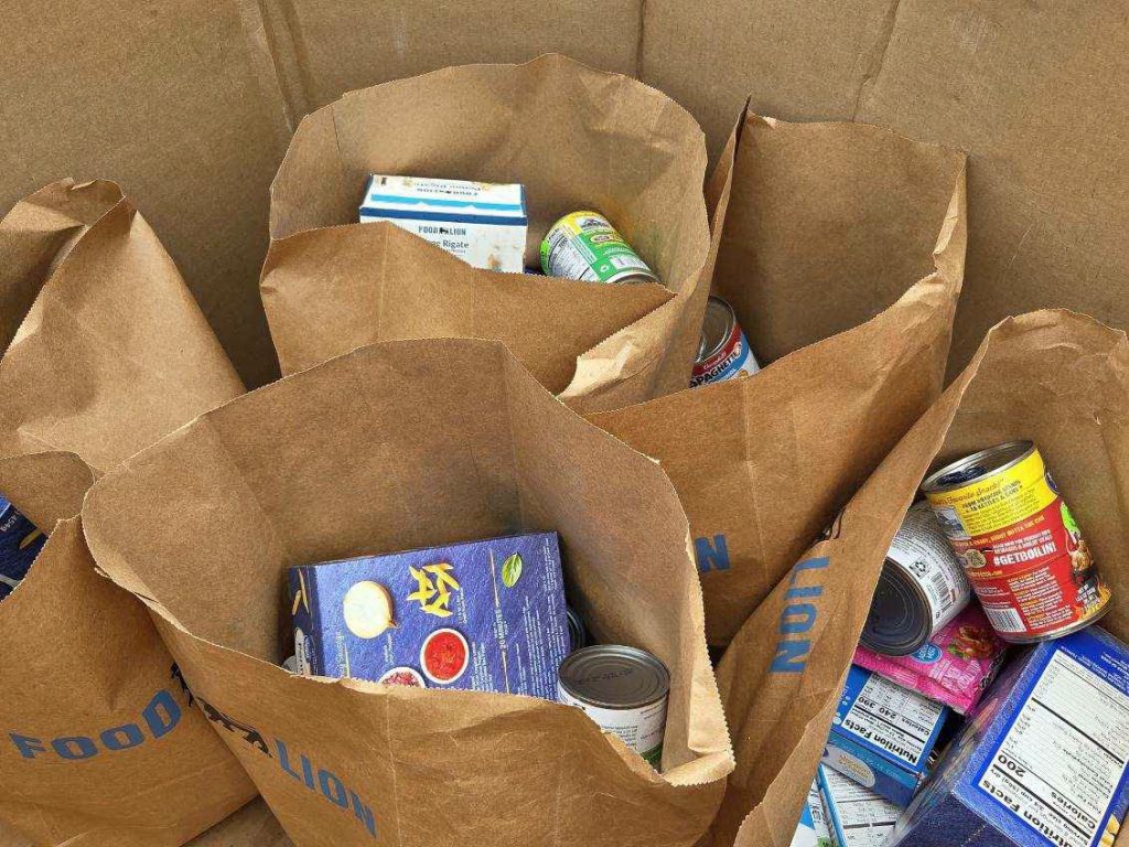 Several brown paper grocery bags filled with canned goods and boxed food items inside a large cardboard box.