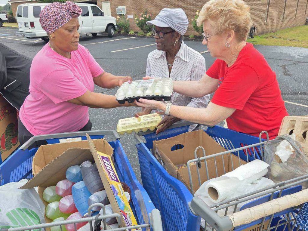 Three women exchanging cartons of eggs outside, standing by shopping carts filled with groceries in a parking lot.