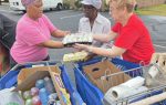 Three women exchanging cartons of eggs outside, standing by shopping carts filled with groceries in a parking lot.