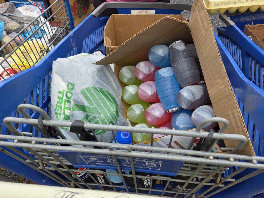 A shopping cart filled with colorful drinks in plastic bottles, a Dollar Tree shopping bag, and various groceries, including eggs and other items in blue baskets.