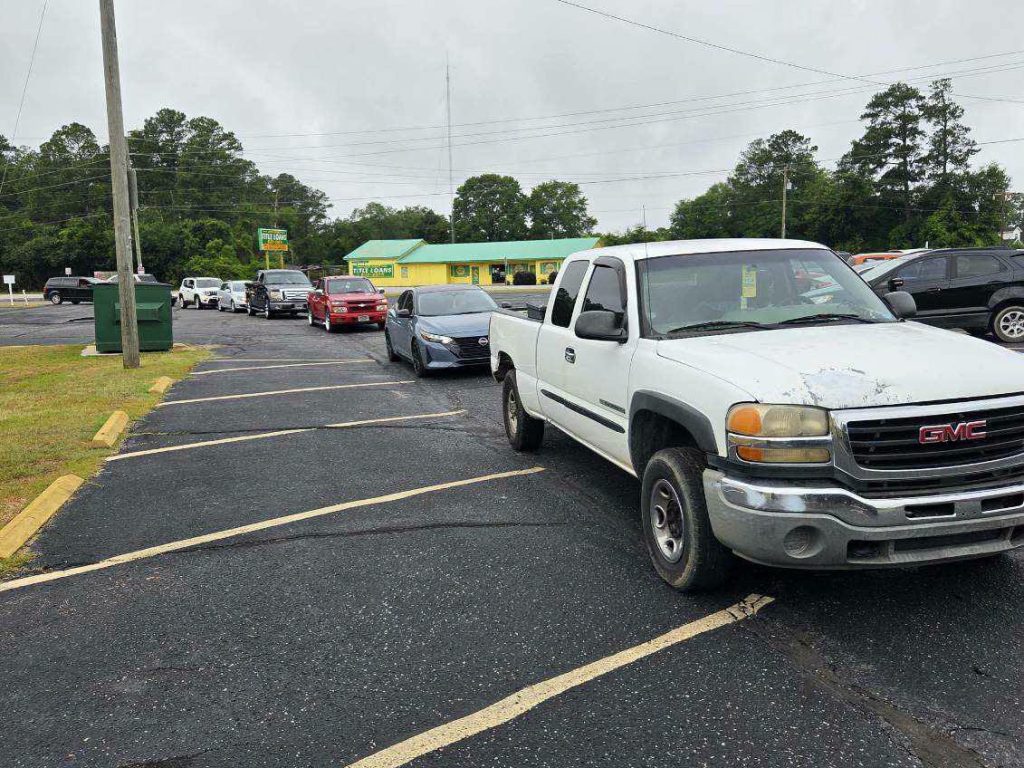 Several cars, including a white GMC pickup, are lined up in a parking lot on an overcast day near a small yellow and green building.