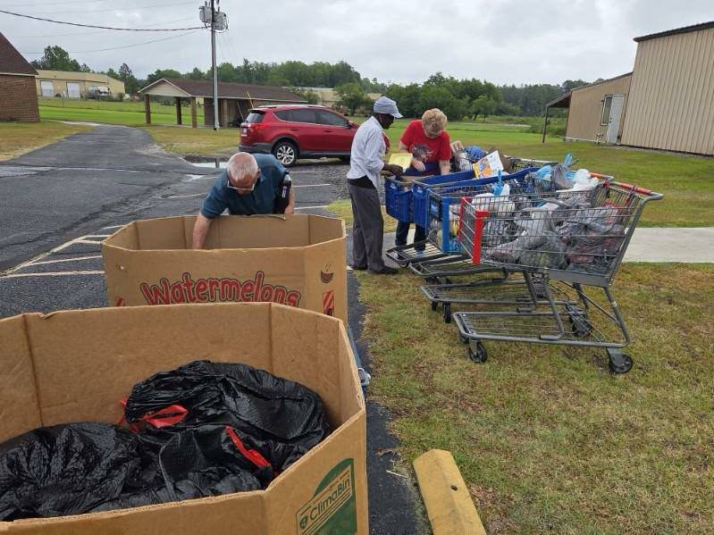 People sorting items from shopping carts into large cardboard bins outdoors, near a parking lot, on an overcast day.