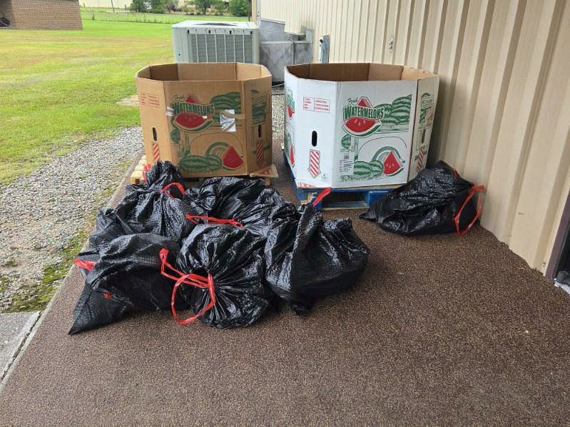 Large watermelon boxes and several black trash bags with red ties outside a building on a brown concrete surface.