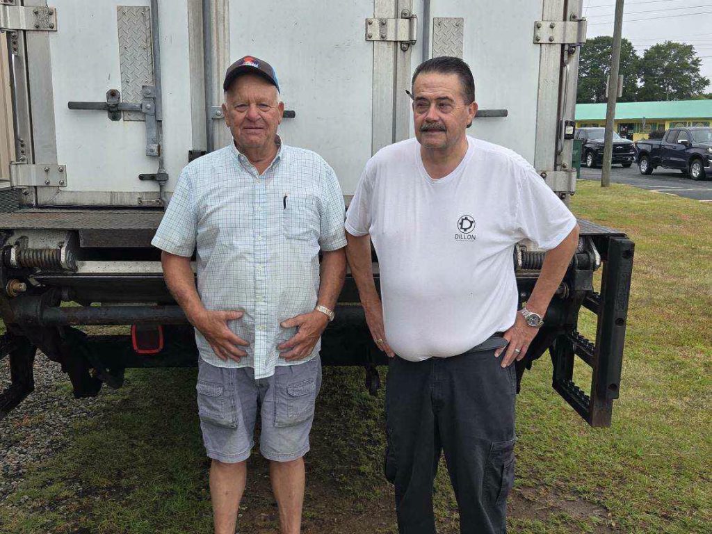 Two older men stand in front of a large truck outdoors, one in a checked shirt and shorts, the other in a white t-shirt and dark pants.