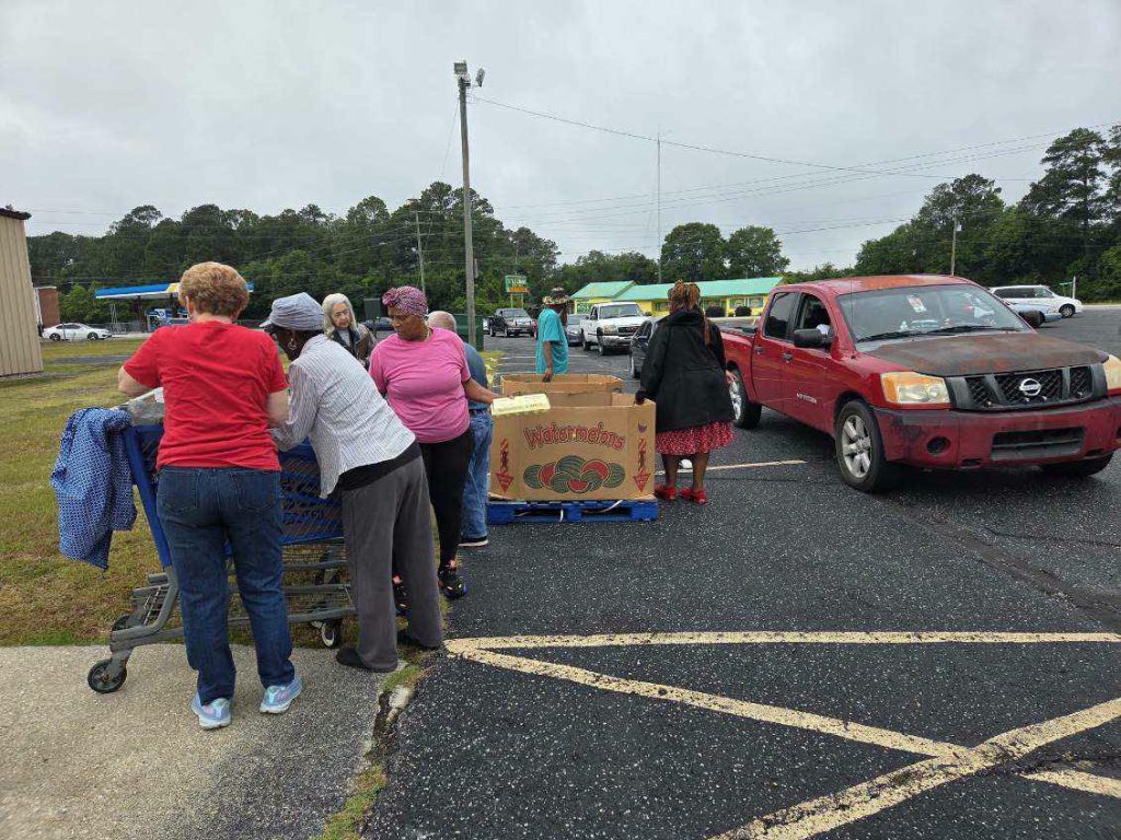 People gather around a large box of watermelons in a parking lot. A red pickup truck and other vehicles are in line nearby.