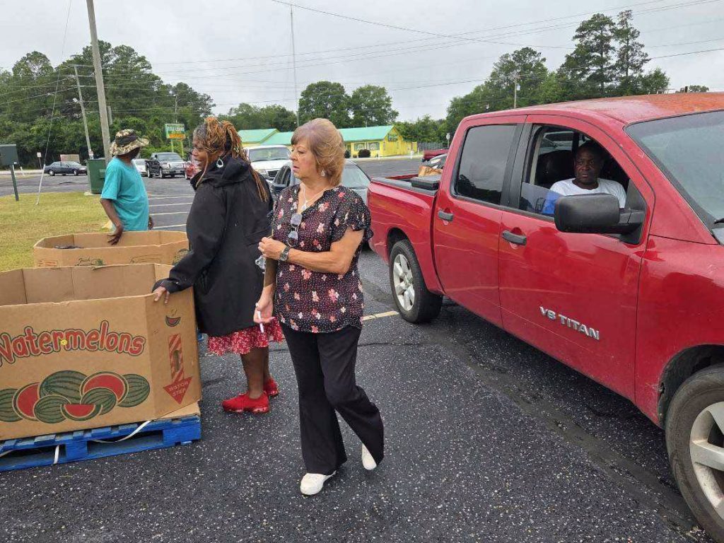 People distribute watermelons outdoors next to large cardboard boxes and a red pickup truck in a parking lot.