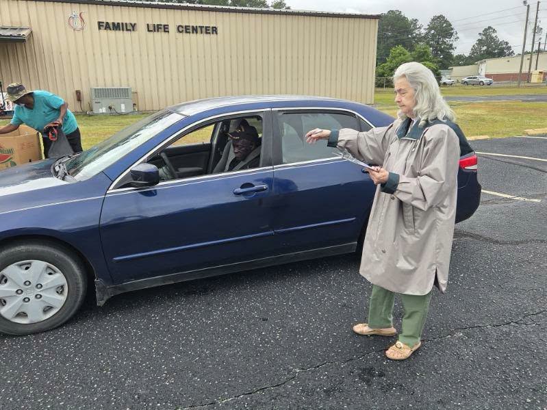 A woman stands beside a blue car speaking to the driver, while another person loads boxes outside the Family Life Center building.