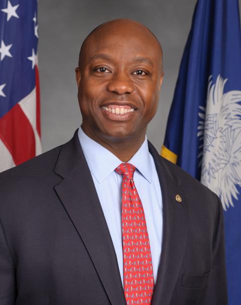 A man in a suit and red tie smiling, standing in front of the American and South Carolina state flags.