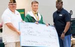 Three men, one in graduation attire, hold a large scholarship check for $1,000 from VFW Post 6091 in a gymnasium.