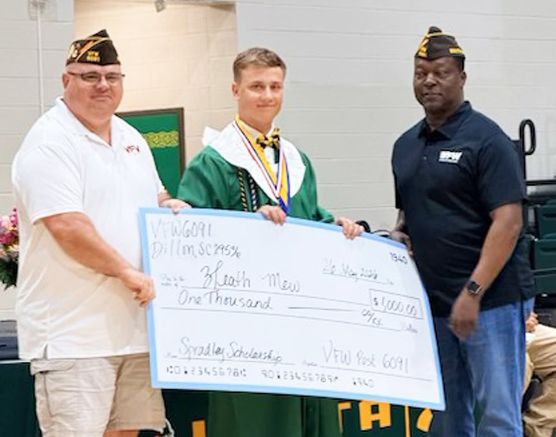 Three men, one in graduation attire, hold a large scholarship check for $1,000 from VFW Post 6091 in a gymnasium.