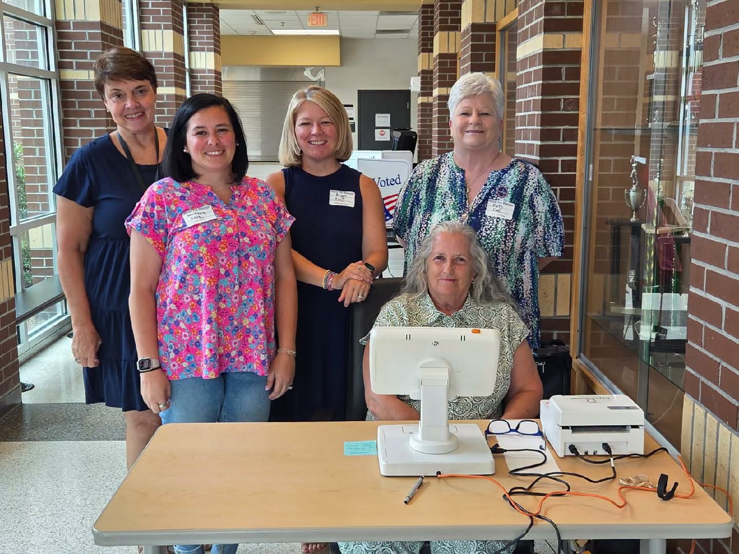Five women, some standing and one seated at a registration table with a computer, inside a building with brick walls and trophy case.