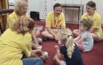 Adults and children sit in a circle on a red carpet, participating in a group activity in a classroom setting. Everyone is wearing yellow shirts except two children.