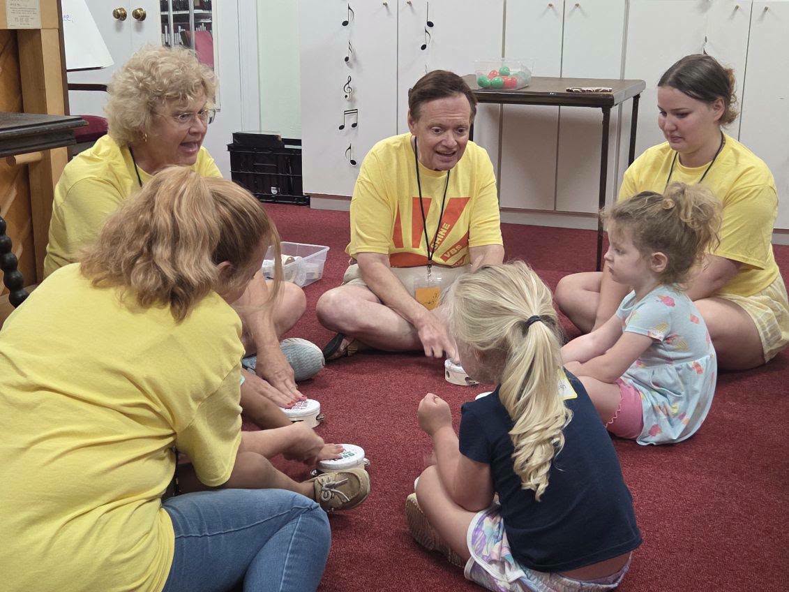 Adults and children sit in a circle on a red carpet, participating in a group activity in a classroom setting. Everyone is wearing yellow shirts except two children.