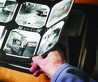 A hand holding open a photo album with black and white photographs, displaying images of people and houses.