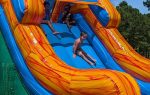 Three children play on a large colorful inflatable water slide outdoors, with one sliding down while two sit at the top, surrounded by green trees.