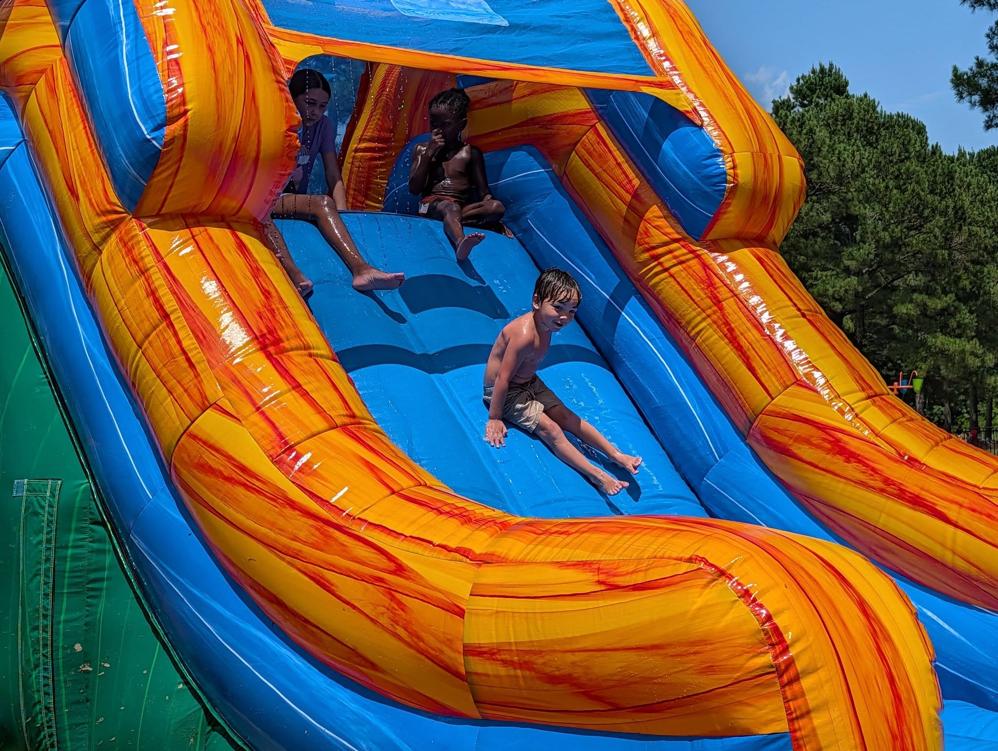 Three children play on a large colorful inflatable water slide outdoors, with one sliding down while two sit at the top, surrounded by green trees.