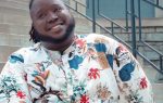 Smiling person in a colorful floral shirt sitting on outdoor steps with metal railings and a brick wall in the background.
