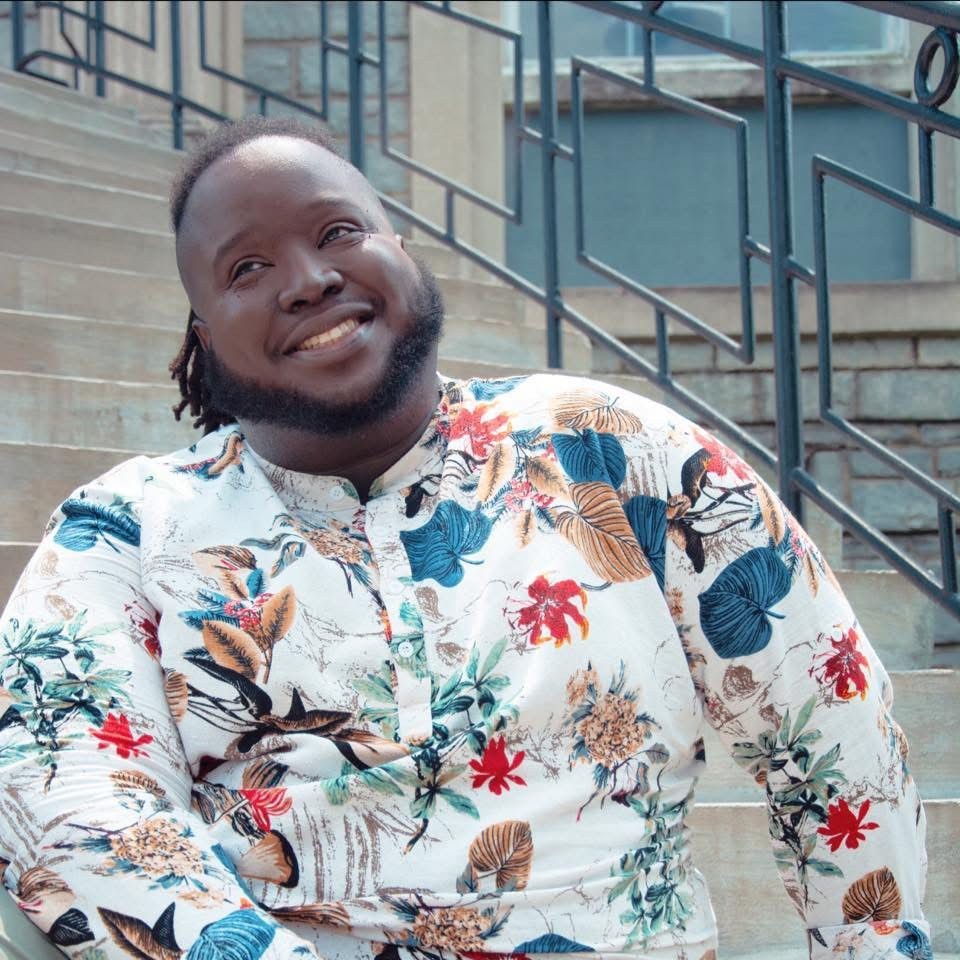 Smiling person in a colorful floral shirt sitting on outdoor steps with metal railings and a brick wall in the background.