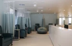 Modern hospital infusion room with reclining chairs, privacy curtains, a bean bag, small tables, and a curved white nurses’ station under bright overhead lights.